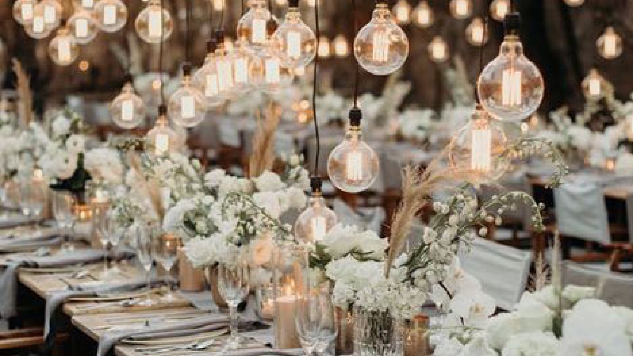 A white themed events table set up for a meal with flowers in the centre and hanging lightbulbs