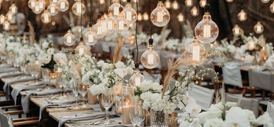 A white themed events table set up for a meal with flowers in the centre and hanging lightbulbs